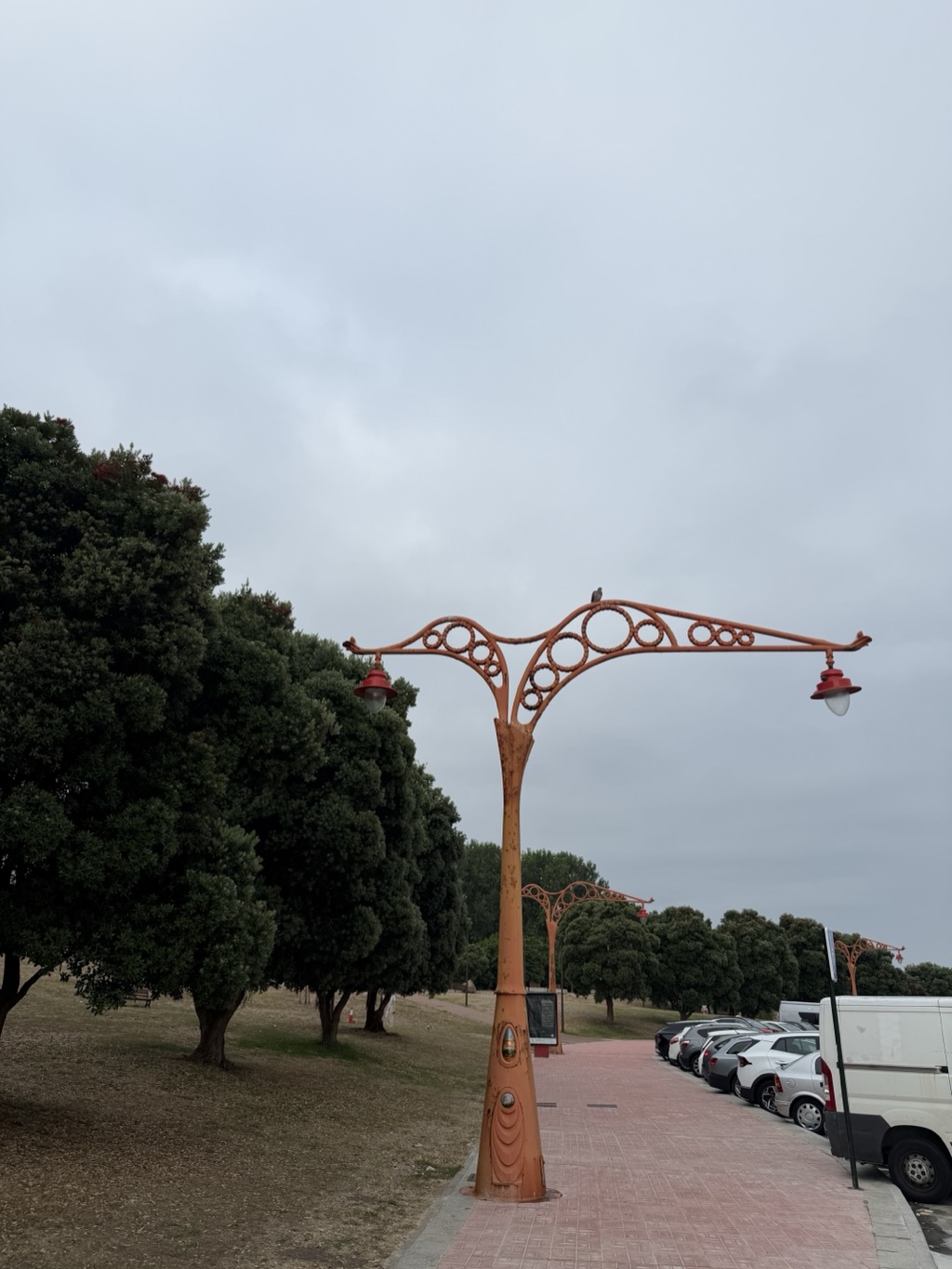 An ornate cast-iron street lamp painted rust orange, standing on a pink brick promenade under a heavy grey overcast sky. Its tapered column is moulded with layered shell-like reliefs at the base and branches at the top into sweeping Art Nouveau arms filled with a lattice of open circles, carrying a red enamelled lantern at each end. A pigeon perches on the crossbar. To the left is parkland with a row of dense dark evergreen trees over dry yellowing grass; to the right a line of parked white and grey cars and a large white van runs along the kerb, with more matching lamps receding into the distance.