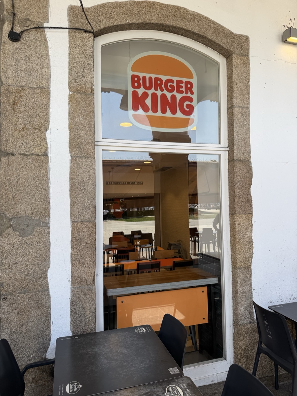 A tall arched window set in a rough grey granite surround with white-painted mortar joints, housing a Burger King. The upper arched pane carries the chain's logo, the red BURGER KING lettering between two halves of an orange bun. Through the lower pane, the empty interior is visible with orange and dark wood tables, black and pale wood chairs, pendant lights and a stone-clad column, and the small line A LA PARRILLA DESDE 1954 on the back wall. Daylight and a stretch of grass and pavement reflect across the glass. In the foreground on the pavement outside are black metal café tables and chairs branded Estrella Galicia, with a QR code sticker on the nearest table top.