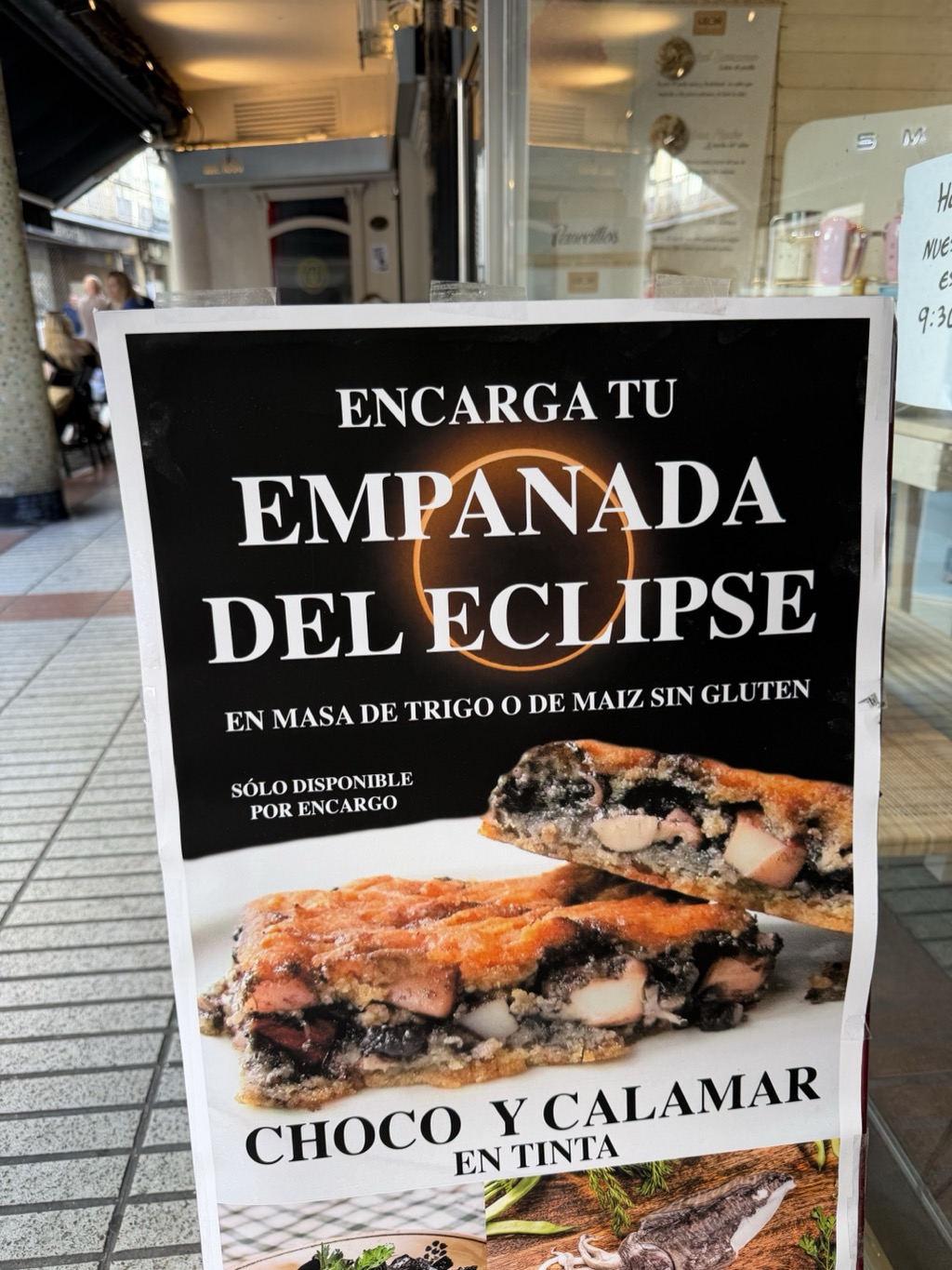 A laminated A-board poster on a tiled pavement outside a café. On a black background, white capitals read ENCARGA TU EMPANADA DEL ECLIPSE, with a glowing orange ring of an eclipsed sun set behind the word EMPANADA. Beneath it, EN MASA DE TRIGO O DE MAIZ SIN GLUTEN and SÓLO DISPONIBLE POR ENCARGO. The lower half shows photographs of a golden lattice-topped pie sliced open to reveal a jet-black squid-ink filling with chunks of white seafood, captioned CHOCO Y CALAMAR EN TINTA, with further images of a plated dish and a whole cuttlefish on a board below. Behind the sign are the café's glass frontage with shelves of jugs and pink mugs, a handwritten notice about 9:30 opening, and a shaded terrace with seated customers further down the street.