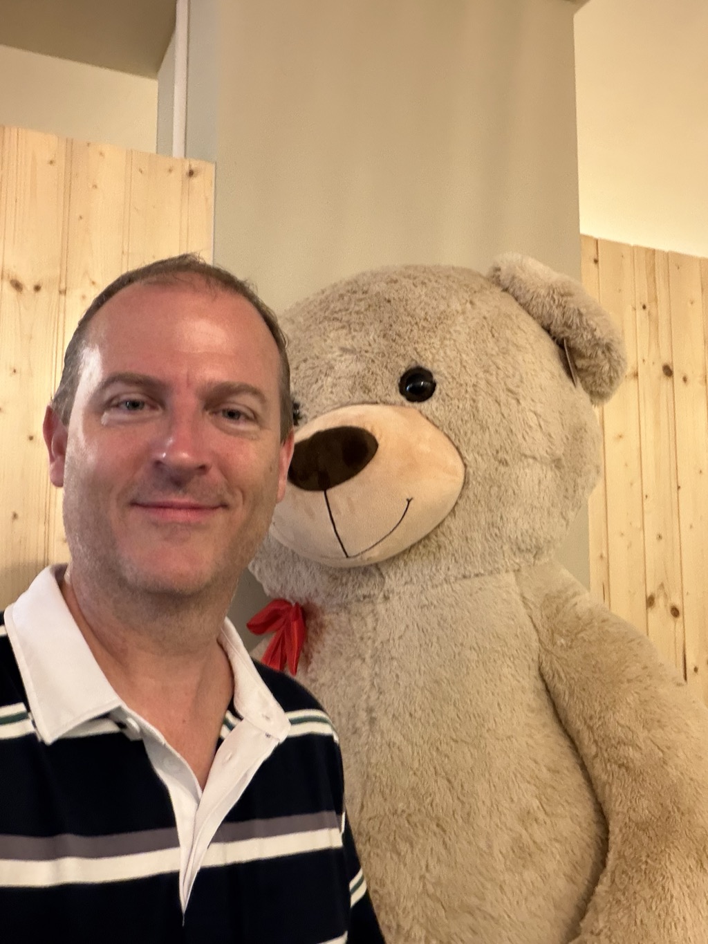A selfie of Brian indoors beside an oversized plush teddy bear that stands taller than he does. He is at the left of the frame with short receding light brown hair, smiling slightly at the camera, wearing a navy rugby shirt with white and grey horizontal stripes and a white collar. The bear fills the right half of the frame, pale beige and fuzzy with a cream muzzle, dark brown nose, stitched smile and a small red ribbon bow at its neck. Behind them are walls of raw pale pine boarding and a cream painted column, lit warmly from a fitting out of shot.