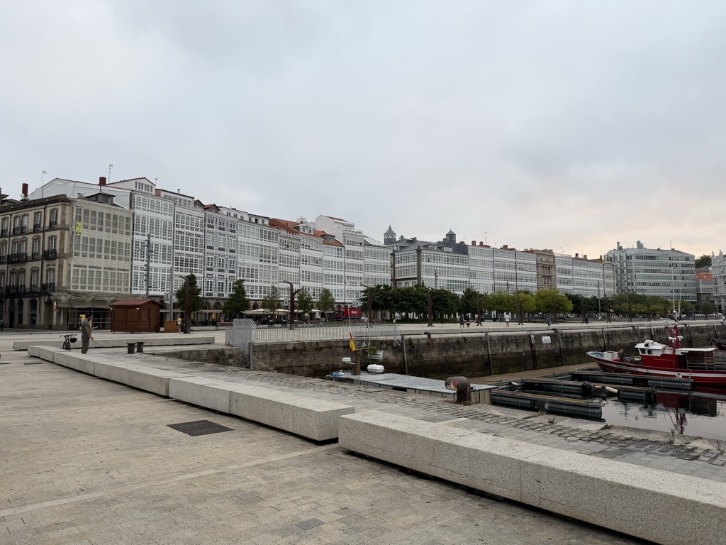 The waterfront of La Coruña seen from an empty granite quayside under a flat grey overcast sky. A long unbroken terrace of tall buildings faces the harbour, their facades entirely covered in the city's characteristic white-painted glazed galleries, tier upon tier of small window panes running the full length of the block. A row of green trees and street lamps lines the promenade in front of them, with a small wooden kiosk and a red vehicle at the left. In the foreground, low granite block benches step down toward the water, where a red and white fishing boat and a line of dark pontoons are moored against the harbour wall. A small Spanish flag flies at the water's edge and one person in a dark jacket walks along the quay.