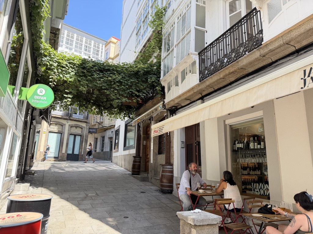 A narrow pedestrian street in bright midday sunshine under a deep blue sky. A dense canopy of green vine leaves is trained across the street on wires about halfway down. The buildings on the right have white-painted glazed galleries stacked above a black wrought-iron balcony and, at ground level, a wine shop with bottles and hanging glasses visible through the window and JAMÓN lettered on the wall. Two people sit at a small red-framed café table on the terrace, a bearded man in a pale shirt leaning forward and a woman in a white top with a glass in front of her, and another woman with dark hair sits at a table in the near right corner. Two large wooden barrels serve as standing tables against the wall. On the left a green OXFAM Intermón projecting sign hangs above a shopfront, and two red drum-shaped tables stand in the near foreground. Further down the sunlit street a person in a white top walks past a row of closed shutters.