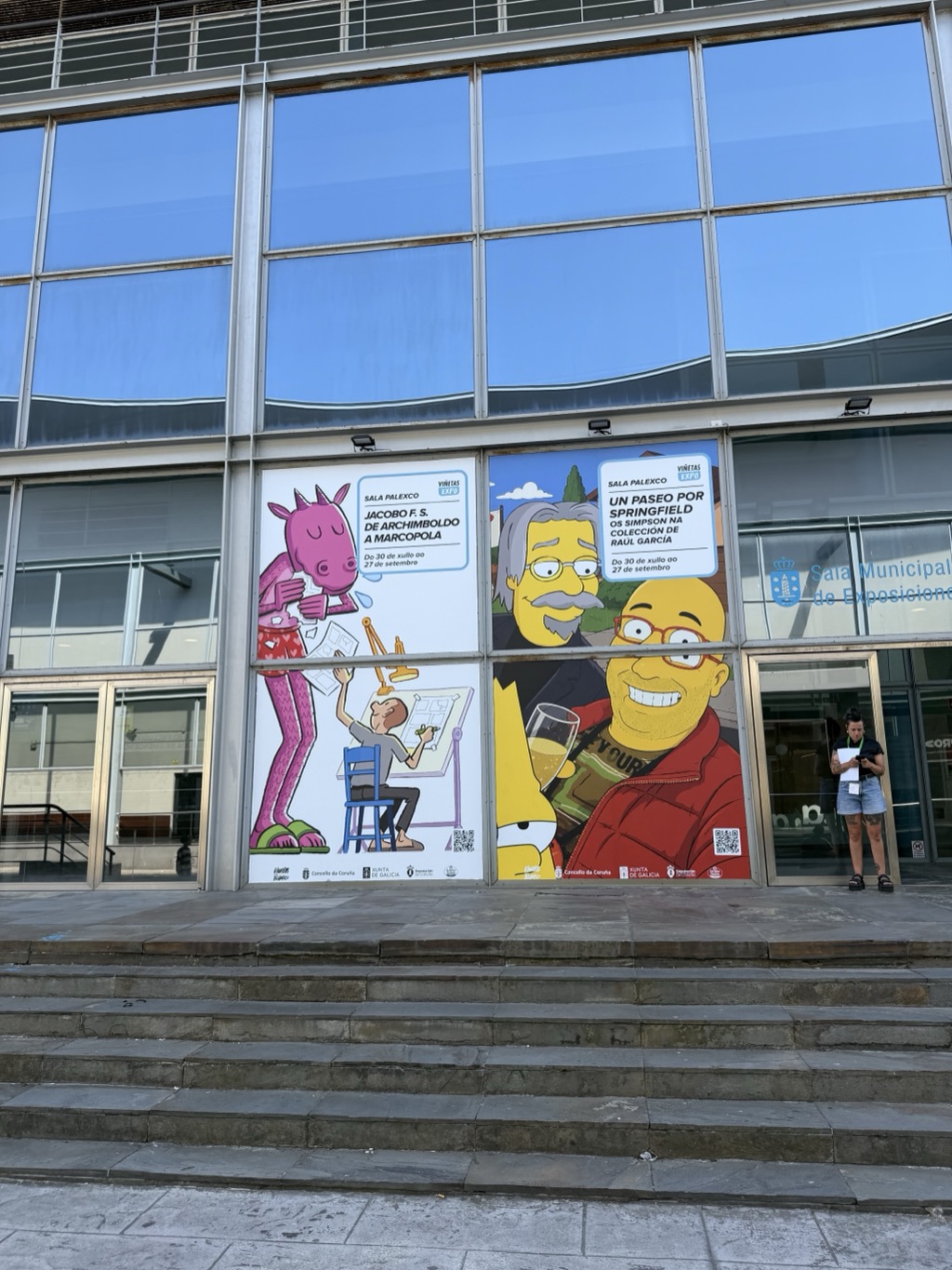 The glass frontage of the Sala Municipal de Exposicións at Palexco, a modern building of blue-tinted mirrored panels in a grey metal grid that reflects a clear blue sky and a wavy white cloud band. Two large exhibition posters are mounted at ground level. The left one, headed SALA PALEXCO VIÑETAS EXPO, reads JACOBO F. S. DE ARCHIMBOLDO A MARCOPOLA and shows a cartoon of a giant pink dragon in red shorts reading papers over the shoulder of a small man drawing at a drafting board. The right one reads UN PASEO POR SPRINGFIELD, OS SIMPSON NA COLECCIÓN DE RAÚL GARCÍA and shows Simpsons-style caricatures of two men, one grey-haired with glasses and a moustache, one bald in a red jacket holding a beer glass, with Bart between them. Both run Do 30 de xullo ao 27 de setembro and carry QR codes and Concello da Coruña and Xunta de Galicia logos. A woman in a black top and denim shorts stands reading a leaflet by the entrance doors at the right, above a broad flight of stone steps.