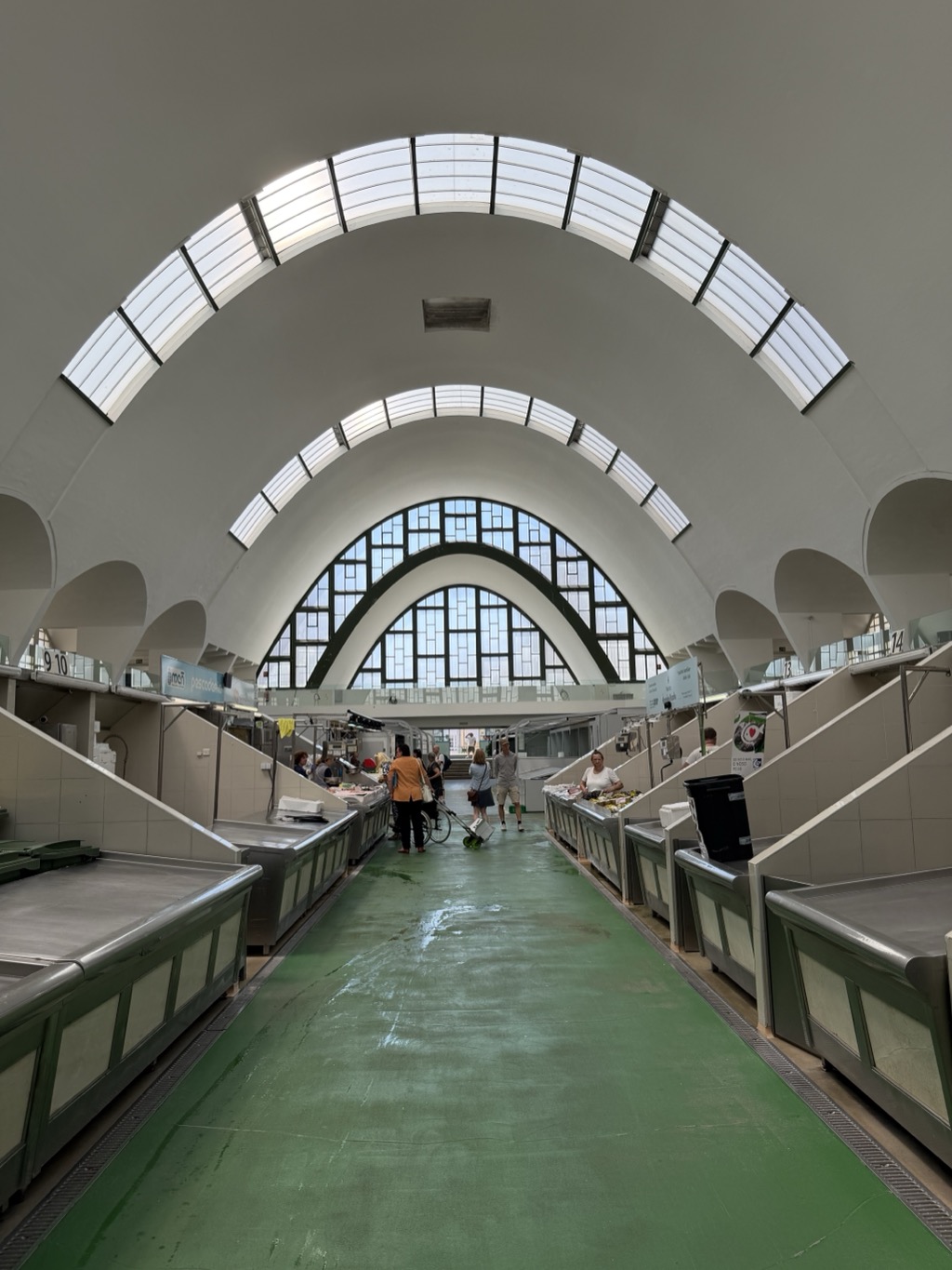 The interior of a covered market hall, looking down a wide central aisle with a green painted floor. A soaring white parabolic concrete vault arches overhead, pierced by two curving bands of skylights, and ends in a tall arched window filled with a grid of small panes in dark green frames, with a second smaller arch inside it. Rows of empty stainless steel and pale green tiled fish counters line both sides, numbered 9, 10, 13 and 14 on hanging signs. Most stalls are cleared and washed down for the day, with a wet patch on the floor and a black wheeled bin at the right. A handful of people are gathered at the far end, including a woman in an orange top with a bicycle and a man in a white shirt behind one of the few counters still holding produce.