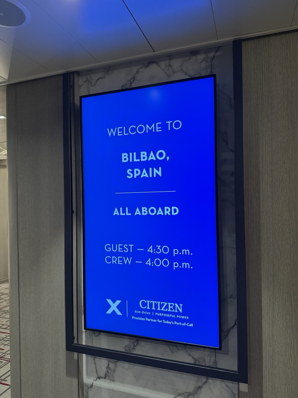 A vertical digital signboard set into a marble and pale timber wall in a ship's lobby, glowing bright blue. White text reads WELCOME TO BILBAO, SPAIN, then below a dividing rule, ALL ABOARD, then GUEST — 4:30 p.m. and CREW — 4:00 p.m. At the bottom is the Celebrity X logo beside CITIZEN, Eco-Drive, PURPOSEFUL POWER and the line Precision Partner for Today's Port-of-Call.