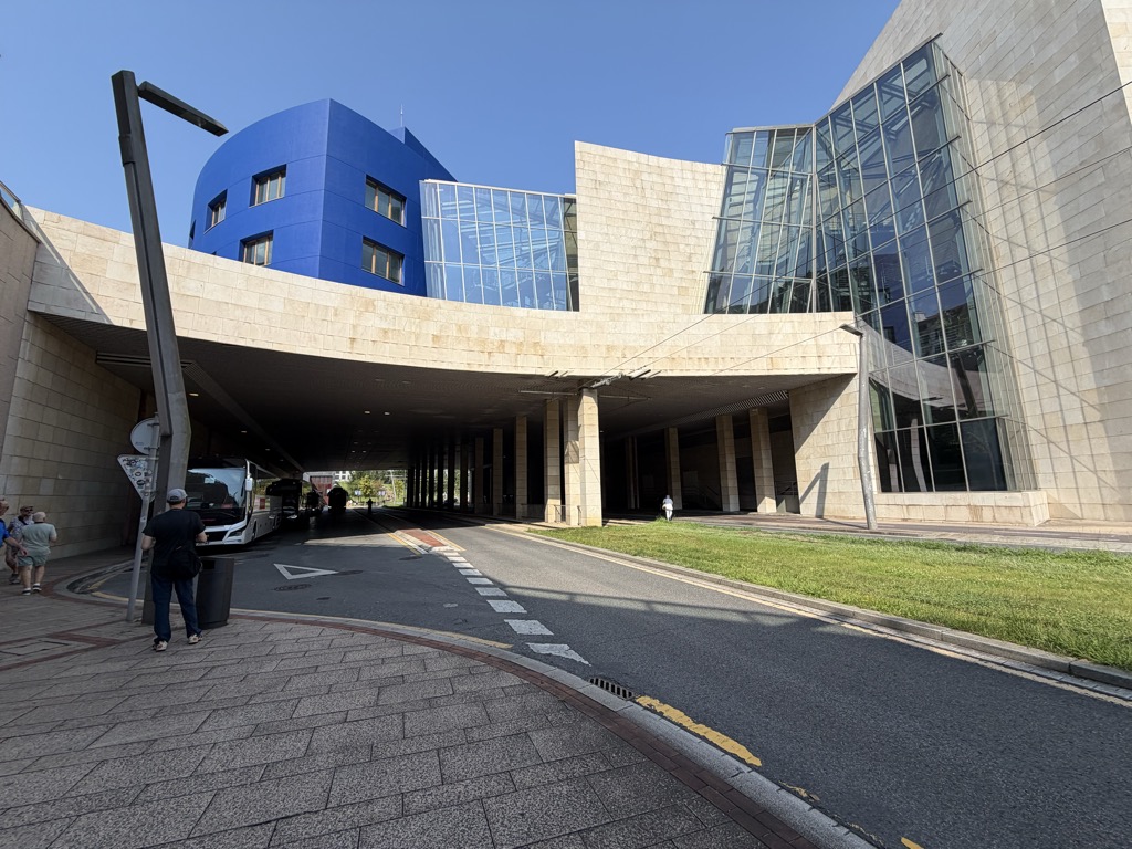 A coach drop-off beneath a large modern building on a bright, cloudless morning. A curving limestone-clad upper storey is carried on square columns over a covered roadway, with a deep blue cylindrical volume rising behind it and a tall angled glass curtain wall to the right. A white coach is parked in the shade under the overhang. A man in a dark t-shirt, jeans and a pale cap walks along the block-paved pavement in the foreground, with two more people at the far left, a strip of bright green lawn to the right and a yellow line painted along the kerb.