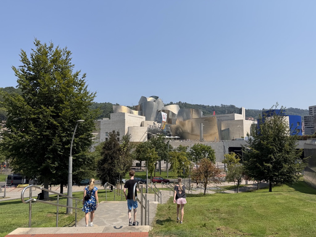 Sherri, Rowan and Siena walk away from the camera down a flight of shallow steps with metal handrails, cut into a grassy slope. Sherri is at the left in a blue and white floral dress, Rowan in the centre in a black t-shirt and blue shorts, and Siena at the right in a black top and pale pink skirt. Below them the road runs past the Guggenheim Museum Bilbao, its titanium curves rising above pale limestone blocks with a banner hanging on the facade. A deep blue office building stands to the right, with young trees, parked cars and green wooded hills behind, under a cloudless blue sky.