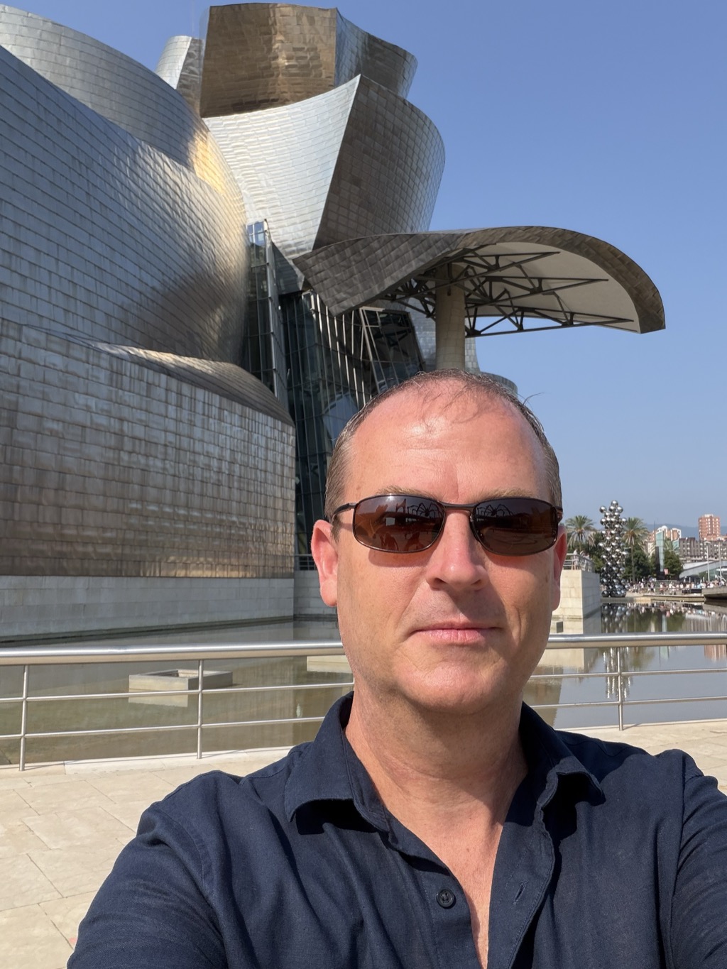 A selfie of Brian in front of the Guggenheim Museum Bilbao, his face filling the lower right of the frame. He wears dark wraparound sunglasses and a navy short-sleeved polo shirt with the collar open. Behind him the museum's titanium panels curve in bands of silver and warm bronze, with the curved metal canopy on its slender column at the right and the glass atrium below. A reflecting pool and steel handrail run behind him, with the mirrored sphere sculpture, palm trees and a bridge visible in the hazy distance under a clear blue sky.