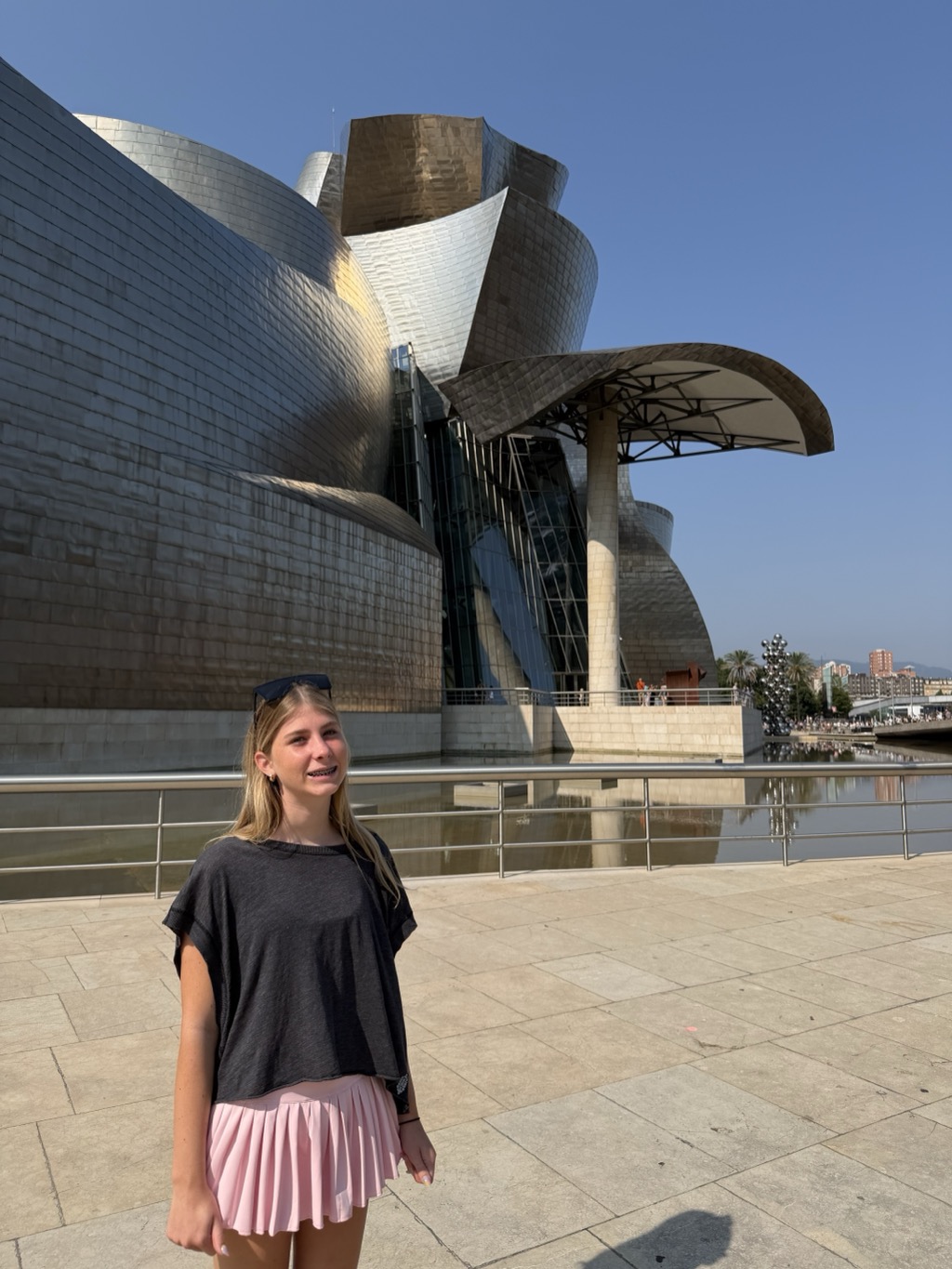 Siena stands smiling on the pale stone terrace in front of the Guggenheim Museum Bilbao, her long blonde hair loose and sunglasses pushed up on her head. She wears a loose charcoal t-shirt over a pale pink pleated skirt. Behind her the museum's titanium-clad volumes twist upward in overlapping silver and bronze curves, with a canopy of curved metal on slender columns to the right and a glass-walled section beneath. A shallow reflecting pool runs along the base of the building, and in the distance a cluster of mirrored spheres, palm trees and apartment blocks are visible under a clear blue sky.