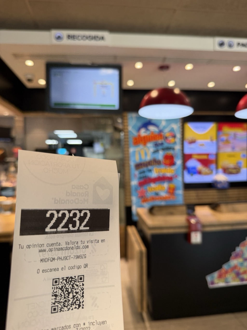 A McDonald's paper order ticket held up in the foreground, printed with the order number 2232 in large white figures on a black band. Smaller Spanish text below invites the customer to rate their visit, above a QR code. Behind it the restaurant counter area is out of focus: a black sign reading RECOGIDA for order collection, an order-status screen on the wall, a row of recessed ceiling spotlights, two deep red pendant lamps, illuminated menu boards and a promotional panel in blue and red.