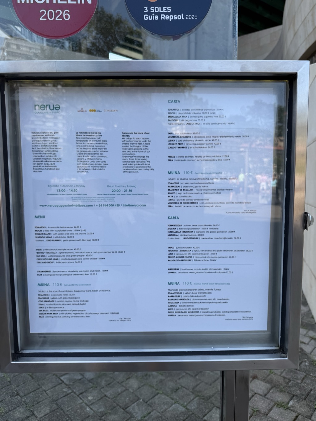 The restaurant's menu behind glass in its stainless steel display case, printed in parallel columns of Basque, Spanish and English. A header block gives the serving hours as 13:00 to 14:30 at midday and 20:00 to 21:30 in the evening, above a paragraph explaining that the kitchen changes its menu three times a year and works with local producers. Individual dishes are listed with prices — tomatoes in aromatic herb sauce, mochi filled with scorpionfish cake, Russian salad with spider crab and red prawns, sea bass, fried suckling lamb, tripe and snout in Biscayan sauce — alongside a tasting menu headed MUINA at 110 €. The MICHELIN 2026 and 3 SOLES Guía Repsol badges are visible at the top of the frame.