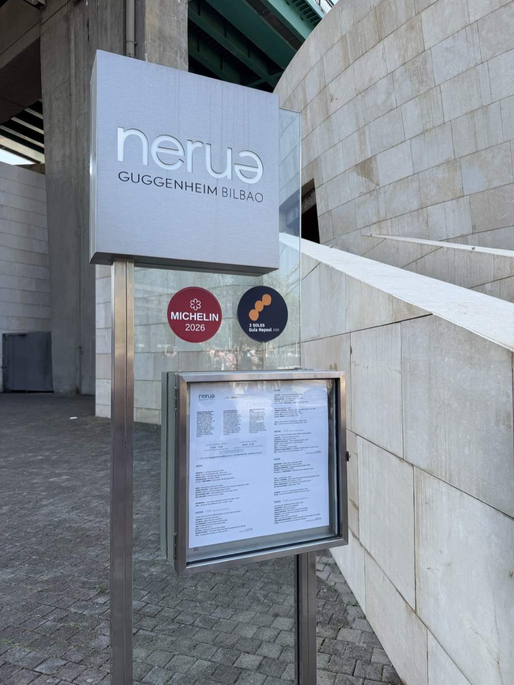 A freestanding stainless steel and glass sign outside a restaurant, lettered nerua in lower-case with GUGGENHEIM BILBAO beneath it. Below the name are two round badges: a dark red MICHELIN 2026 disc with the star emblem, and a navy 3 SOLES Guía Repsol disc with three orange dots. A lockable display case mounted below holds the printed menu. Behind the sign, the museum's pale limestone wall curves away up a flight of steps, and the green-painted steel underside of a road bridge crosses overhead.