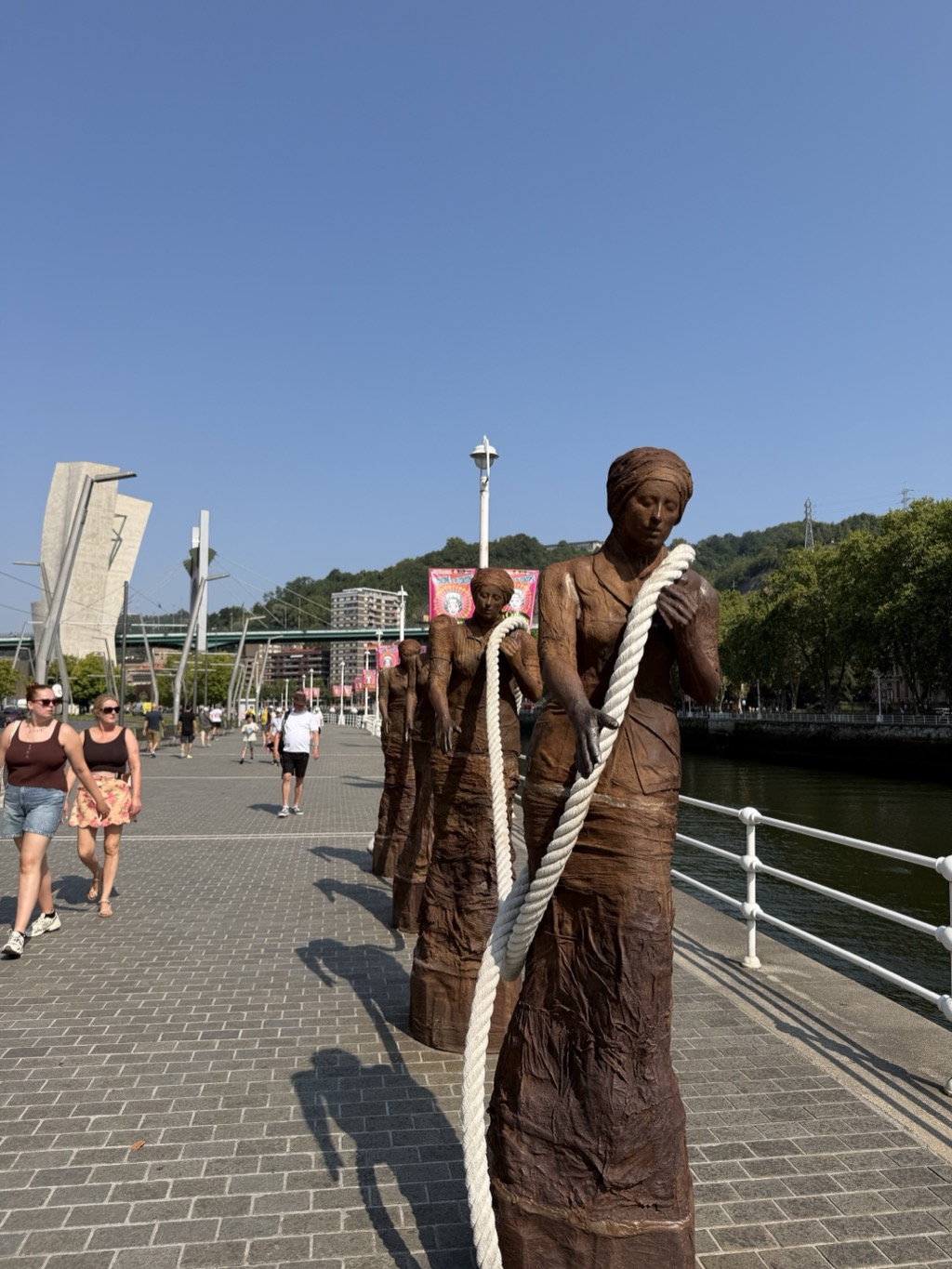 A row of life-size rust-brown sculpted female figures strung out along a riverside promenade, each leaning into a thick white rope that runs from the front figure back through the line, as though hauling a barge upstream. The nearest figure is in a long dress with a cloth wound round her head, her hands gripping the rope. Two women in vest tops and shorts walk past on the left and more pedestrians are further along the paving. A white-painted railing runs beside the green river to the right, with plane trees, a road bridge on slender masts, banners on lampposts and a wooded hillside beyond, all under a clear blue sky.