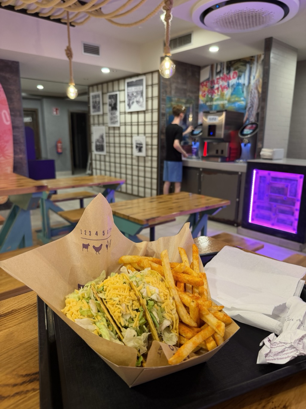A cardboard carton on a black tray holding two crunchy tacos filled with shredded lettuce and grated yellow cheese, alongside a pile of orange seasoned fries and a folded paper napkin. The carton is printed with a row of numbers and small animal silhouettes. Behind the tray the restaurant interior recedes: wooden picnic-style tables with brightly painted legs in teal and purple, pendant bulbs hung on lengths of rope, a grid-patterned partition, framed black and white photographs, and a self-serve drinks machine lit with purple neon where a teenager in a black t-shirt and blue shorts is filling a cup.