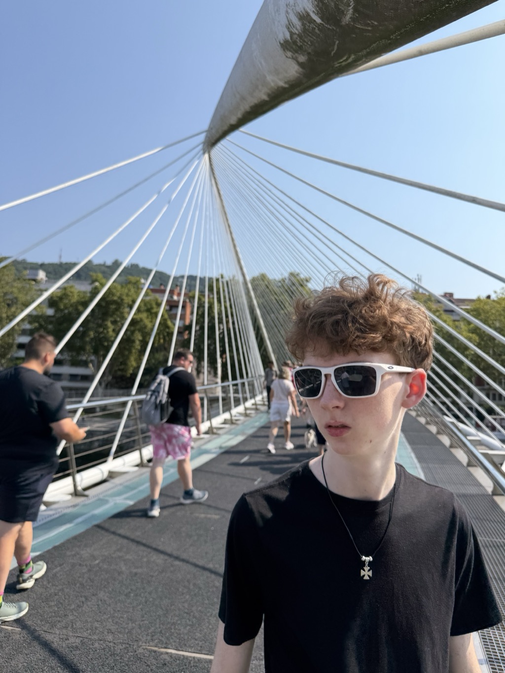 Rowan stands on the Zubizuri footbridge over the river in Bilbao, looking toward the camera in white-framed sunglasses and a black t-shirt, with a silver cross pendant on a black cord. His curly reddish-brown hair catches the sun. Behind him the bridge's white steel arch sweeps overhead and its cable stays fan down to the deck on both sides. The walking surface runs away in dark asphalt with a pale turquoise strip of the original glass decking along the left edge. Several other pedestrians walk ahead, including a man with a grey backpack in pink patterned shorts, with green trees and apartment buildings on the far bank.