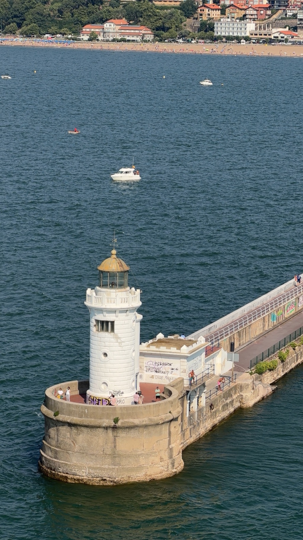 The Faro del Rompeolas, a white stone lighthouse at the end of the long Getxo breakwater, seen from a departing ship. The round tower has a ring of small square windows, a railed gallery with a stone balustrade, and a glazed lantern room capped by a green-bronze dome and a weathervane. It stands on a massive semicircular stone bastion whose lower courses are stained green at the waterline. Graffiti in coloured spray paint covers the base of the tower and the small flat-roofed building beside it. A handful of people stand on the bastion and along the mole, which runs away to the right behind a white railing. Two small motorboats and a rowing skiff are on the blue water, and behind them a crowded sandy beach curves along the shore below red-roofed villas and a wooded headland.