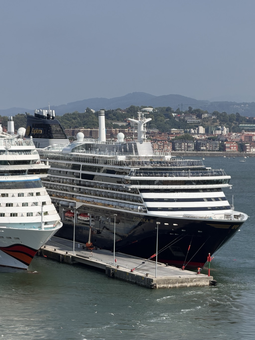 Two cruise ships lie alongside a concrete quay, seen from high on a third ship. In the foreground the dark blue hull and white superstructure of EXPLORA III fills the frame, her name lettered on the bow above a gold band, with mooring lines running down to bollards on the pier and lifeboats slung along her side. Beyond her bow is another ship of the same line, her funnel lettered Explora in gold on black and white stripes. At the left edge a third vessel shows a white hull with a red and orange swoosh. Behind the ships, a town of red-roofed apartment blocks and villas climbs a green hillside, with hazy blue mountains beyond and small yachts moored on the water.