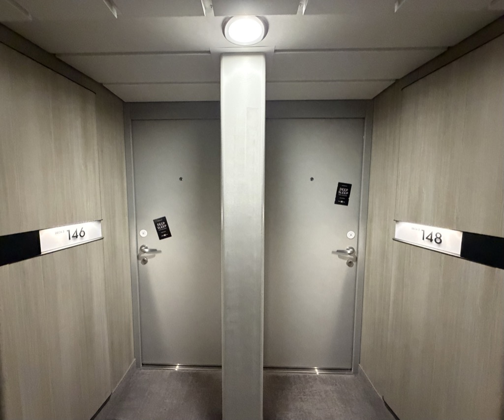 A short alcove off a ship's corridor with two stateroom doors side by side, separated by a square structural column. The doors are pale grey with brushed steel lever handles and card readers, and each carries a small black door hanger printed DEEP SLEEP in white. Cream vertical-grained timber panelling lines the alcove walls, with a black band at waist height carrying white cabin number plates reading 146 on the left and 148 on the right, each with DECK 3 in small type above. A single round ceiling light glows overhead and grey carpet runs across the threshold.