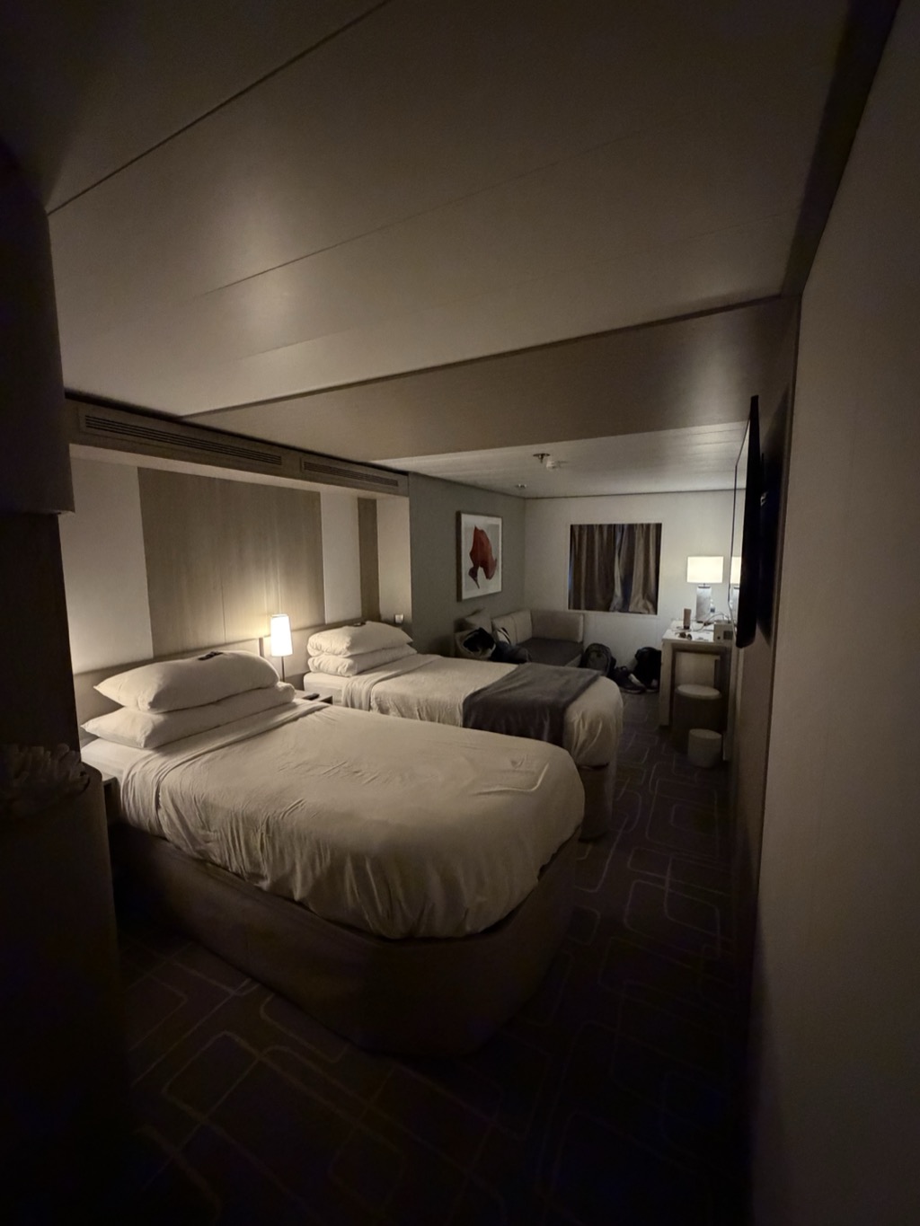 A dimly lit cruise ship stateroom at night, photographed from the entry passage. Two single beds made up in cream bedding stand side by side in the foreground, the nearer one with a folded grey throw across its foot and stacked white pillows against a pale timber headboard wall. A single bedside lamp is on between them. Beyond the beds a grey sofa under a framed picture of a red fish holds a jumble of packed bags, with more luggage on the floor beside it. A curtained window, a writing desk with a lit lamp and a wall-mounted television fill the far corner, and the floor is laid with a dark patterned carpet.