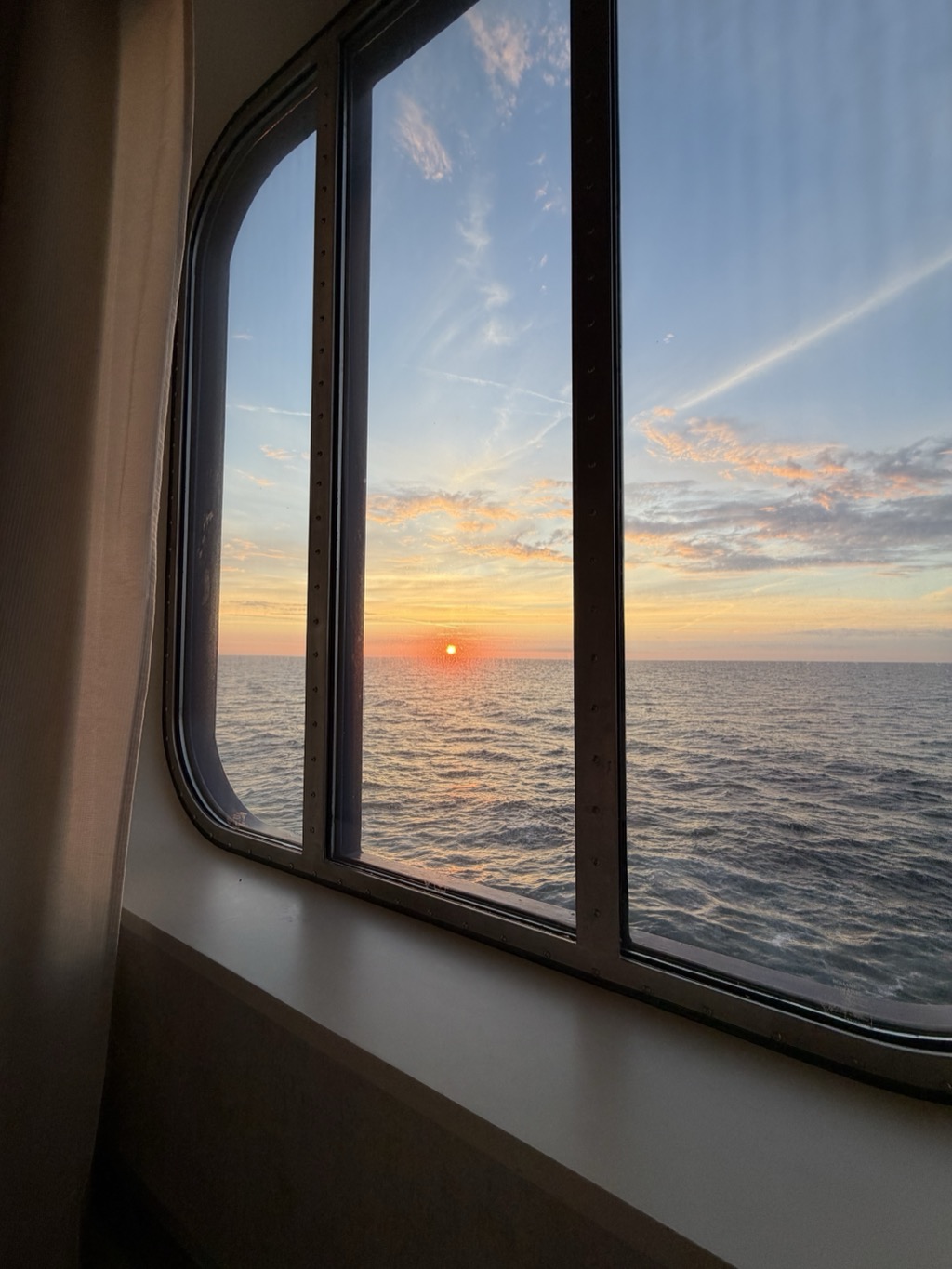A sunset over open ocean seen from inside the ship through a large window divided into panes by dark metal mullions studded with rivets. The sun sits as a small bright disc just above a flat, clean horizon, laying a pale gold path across gently rippled deep blue water. Above it the sky grades from warm apricot at the horizon through pale gold to soft blue, streaked with thin cirrus and two long contrails, with a band of small pink and grey clouds catching the light to the right. A cream curtain hangs at the left edge of the frame and the pale windowsill runs across the bottom.