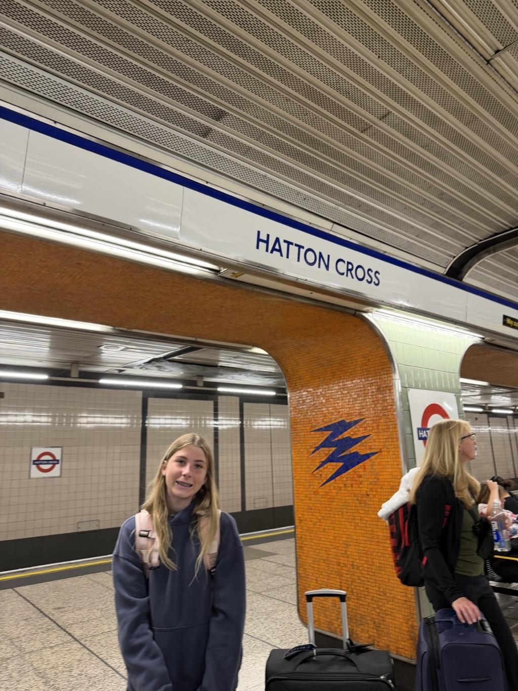 Siena stands on the Underground platform at Hatton Cross, smiling toward the camera with her long blonde hair down, wearing a slate blue hoodie and a pale pink backpack. Behind her a white sign lettered HATTON CROSS in blue runs along the tunnel wall beneath a ribbed metal ceiling, and a burnt orange tiled pillar carries the station's blue zigzag motif. A roundel is mounted on the cream tiled wall of the opposite platform across the track. To the right, a woman with blonde hair and glasses in a black jacket stands with a rucksack and a water bottle, and wheeled suitcases are lined up along the platform edge in the foreground.