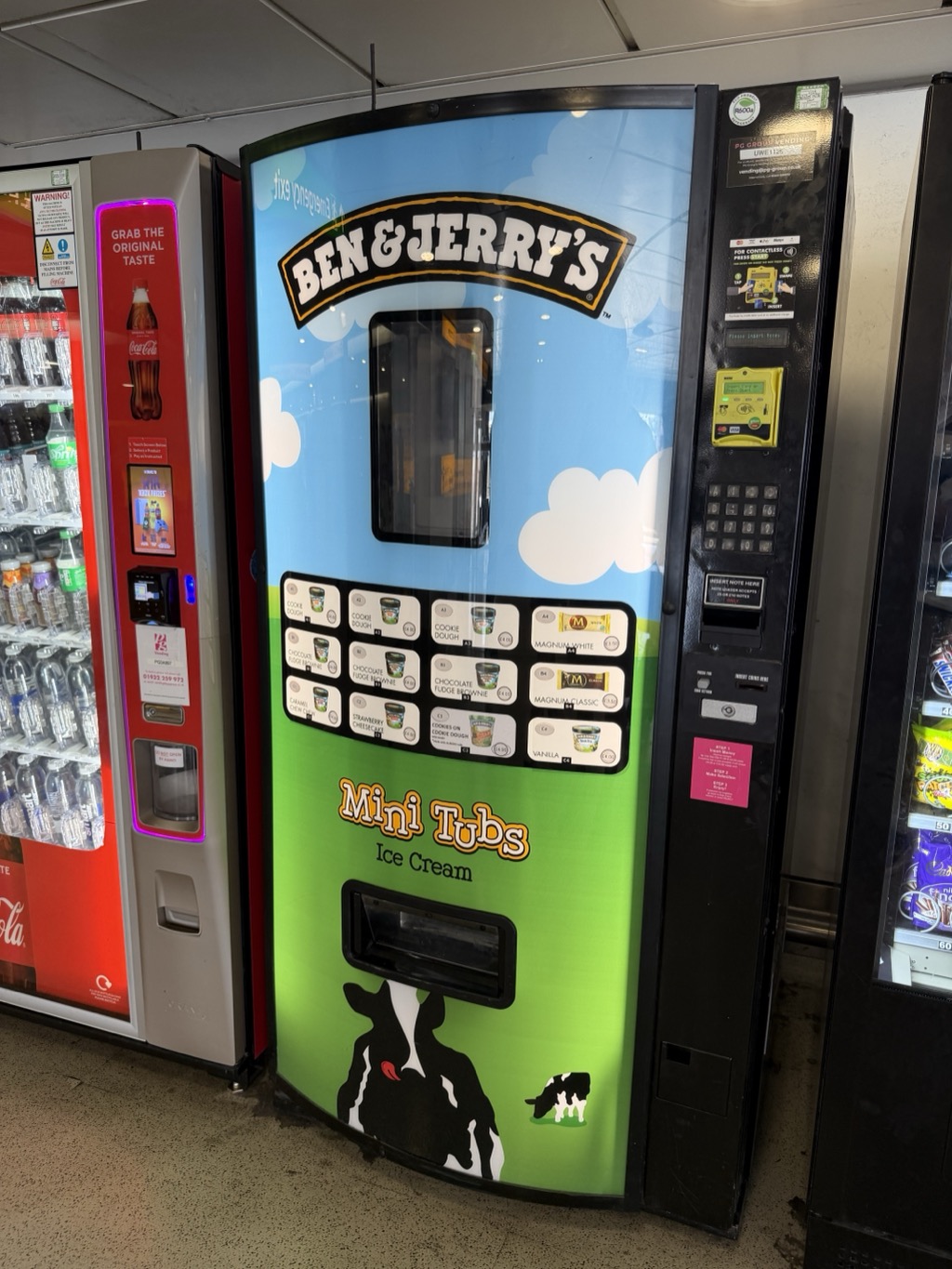 A Ben & Jerry's ice cream vending machine in an airport concourse, its curved front wrapped in a blue sky and green field design with a black and white cow at the bottom and the words Mini Tubs, Ice Cream in yellow lettering. A grid of product buttons shows mini tubs of Cookie Dough, Chocolate Fudge Brownie, Caramel Chew Chew, Strawberry Cheesecake and Vanilla alongside Magnum White and Magnum Classic bars, with prices beside each. A black payment column at the right carries a contactless reader, a keypad, a note slot and a coin slot. A red Coca-Cola drinks machine stands to the left beside a chilled cabinet of bottled water, and a snack machine is partly visible at the right.