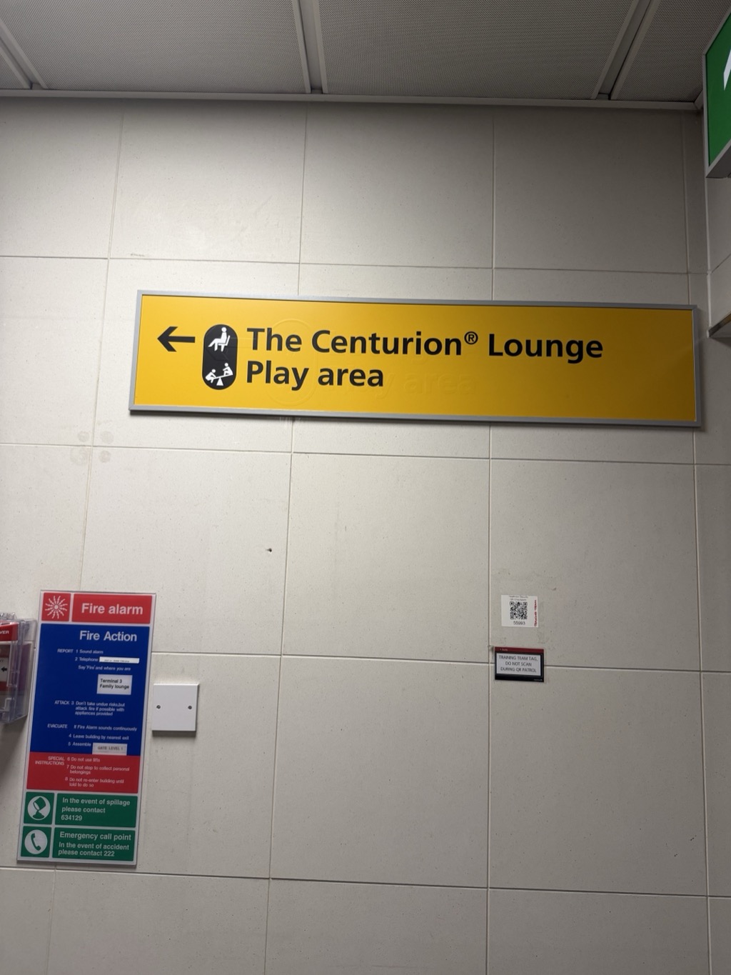 A yellow airport wayfinding sign mounted on a white tiled wall, lettered in black with a left-pointing arrow and two pictograms — a seated figure and an adult with a child on a slide. The text reads The Centurion® Lounge and, on a second line, Play area. Below and to the left, a Fire Action notice in red, blue and green panels gives numbered emergency instructions for the Terminal 3 Family Lounge, along with a spillage contact number and an emergency call point extension. A small QR code label to the right is captioned as a training team tag not to be scanned during patrol, and the corner of a green emergency exit sign shows at the top right.