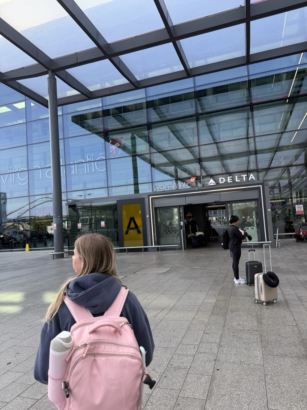 Siena, seen from behind with her pink backpack over a navy hoodie, walks toward the entrance of Heathrow Terminal 3 under a glass canopy on a bright morning. The terminal's glazed facade carries the Virgin Atlantic name and logo in white across the glass, with a smaller Virgin Atlantic and DELTA sign above the automatic doors. A yellow panel to the left of the doors is marked with a large letter A. A woman in black activewear stands to the right beside a black cabin bag and a beige suitcase with a travel pillow looped over the handle, and other travellers with luggage are visible through the doors.