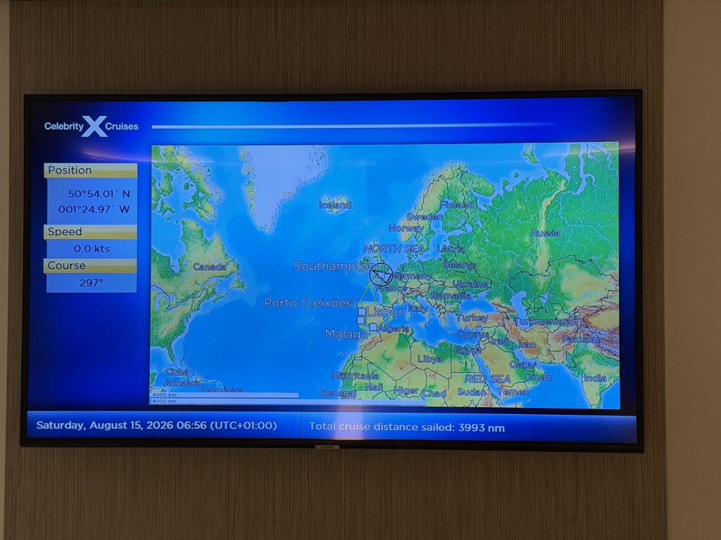 A wall-mounted flat-screen television in a wood-panelled stateroom, showing the ship's navigation channel. A Celebrity X Cruises logo sits at the top left above a world map centred on the Atlantic, with Europe, Africa and the eastern seaboard of North America labelled. A small ship symbol is circled at Southampton, and the ports of Porto (Leixões), Lisbon and Málaga are marked down the Iberian coast. A panel at the left lists Position 50°54.01′ N, 001°24.97′ W, Speed 0.0 kts and Course 297°. A blue bar across the bottom reads Saturday, August 15, 2026 06:56 (UTC+01:00) and Total cruise distance sailed: 3993 nm.